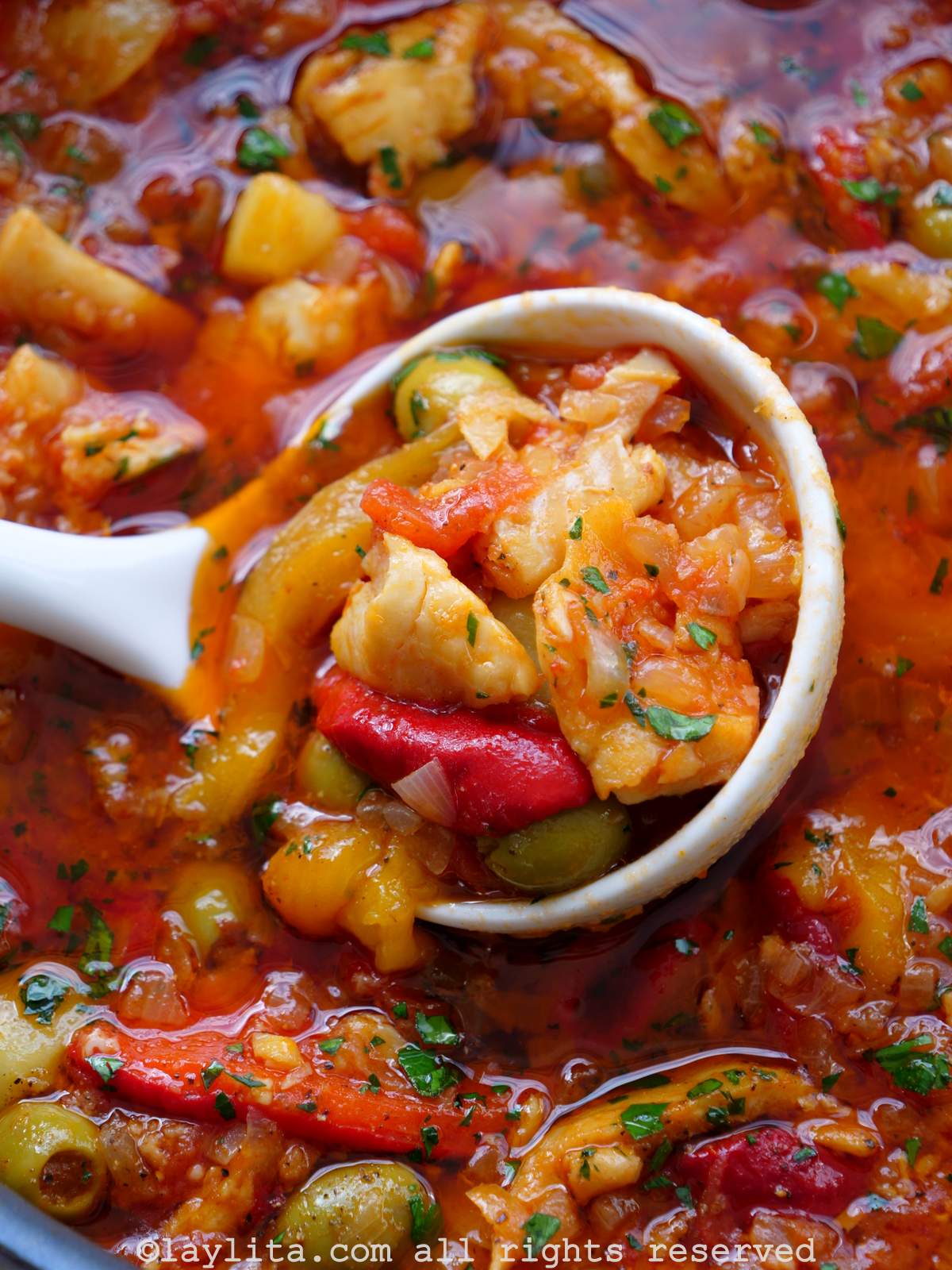 Close up of salted cod fish in a tomato and roasted pepper sauce with a ladle scooping up a portion from the pot.