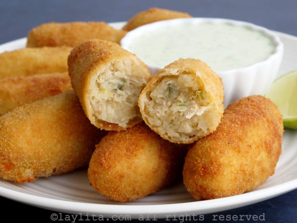 Oval-shaped tuna potato croquettes on a plate served with a side of sauce with one croquette broken in half to show the inside.
