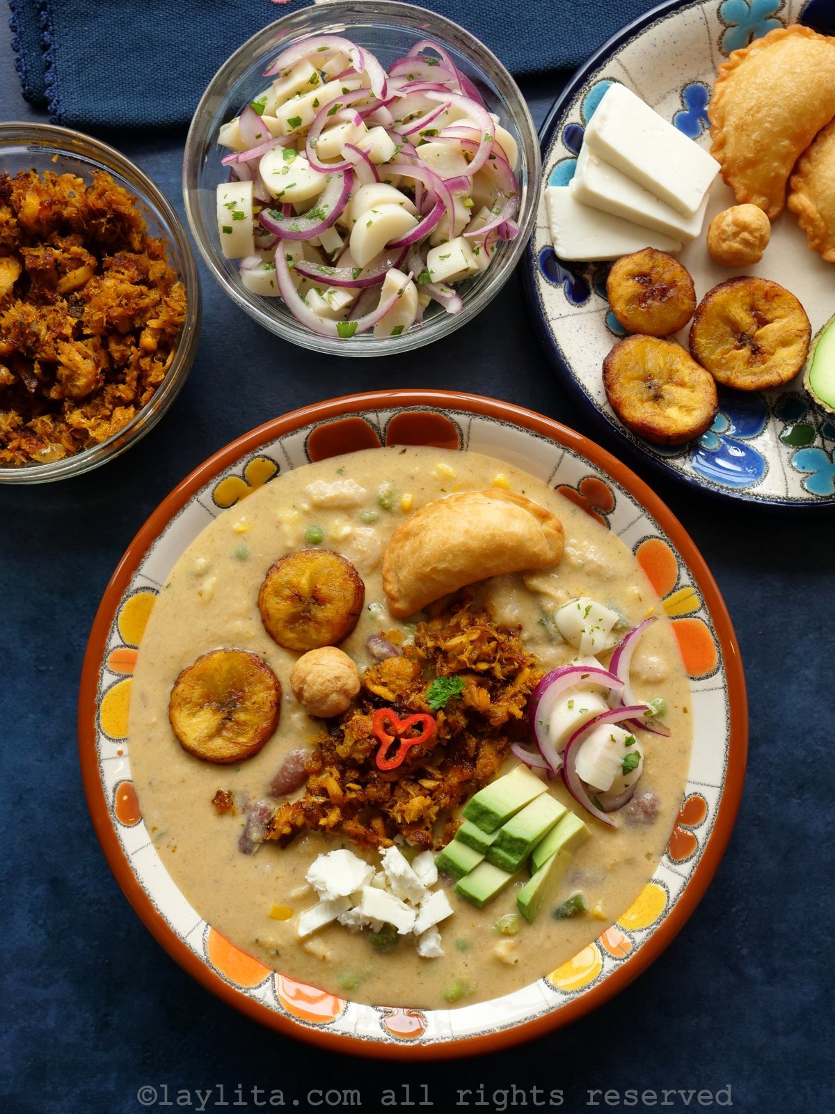 Fanesca de atun in bowl topped with empanadas, fried plantains, avocado, cheese and other garnishes. Additional garnishes in the background.
