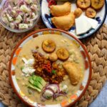 A bowl of Ecuadorian fanesca and side dishes include heart of palm salad, fried plantains, empanadas, and avocados on a rattan placemat