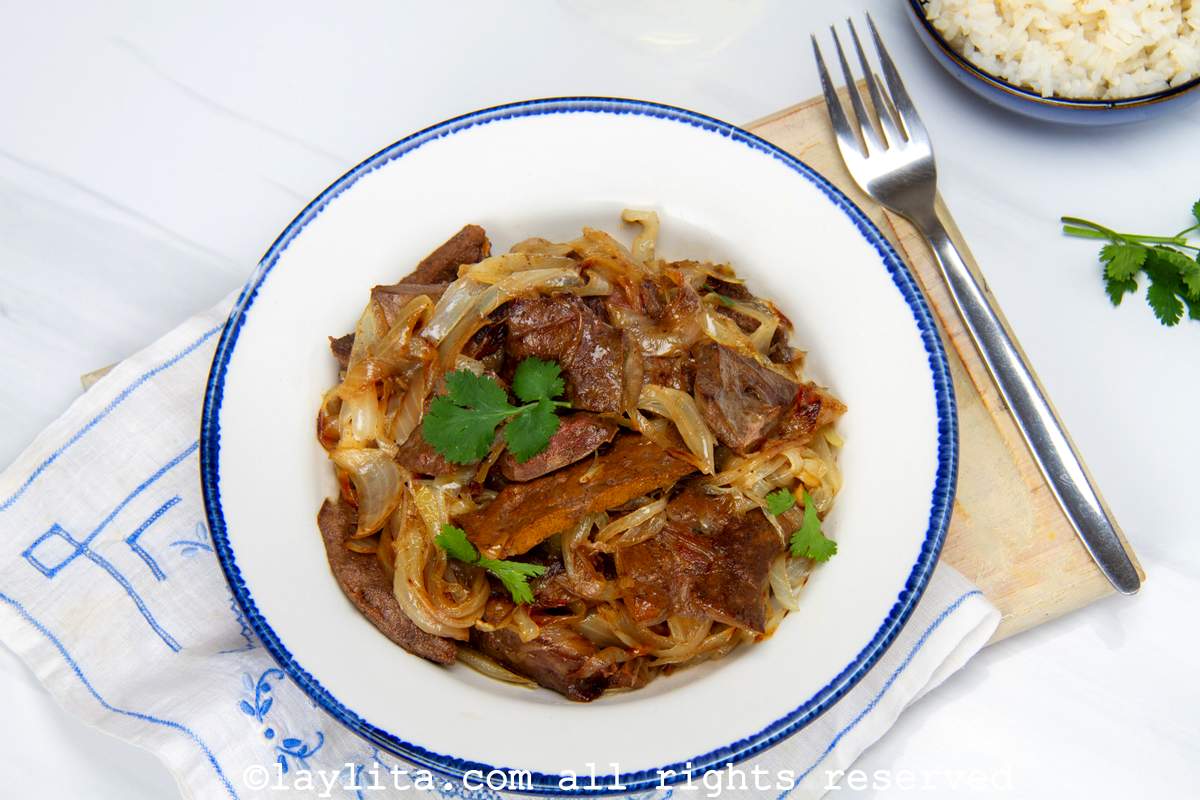 Bowl of sauteed liver with onions served next to a fork on a wooden board