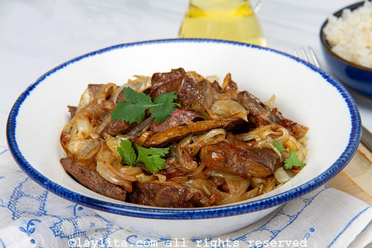 Bowl of liver with onions or hígado encebollado on a wooden board lined with a linen next to a bowl of rice.