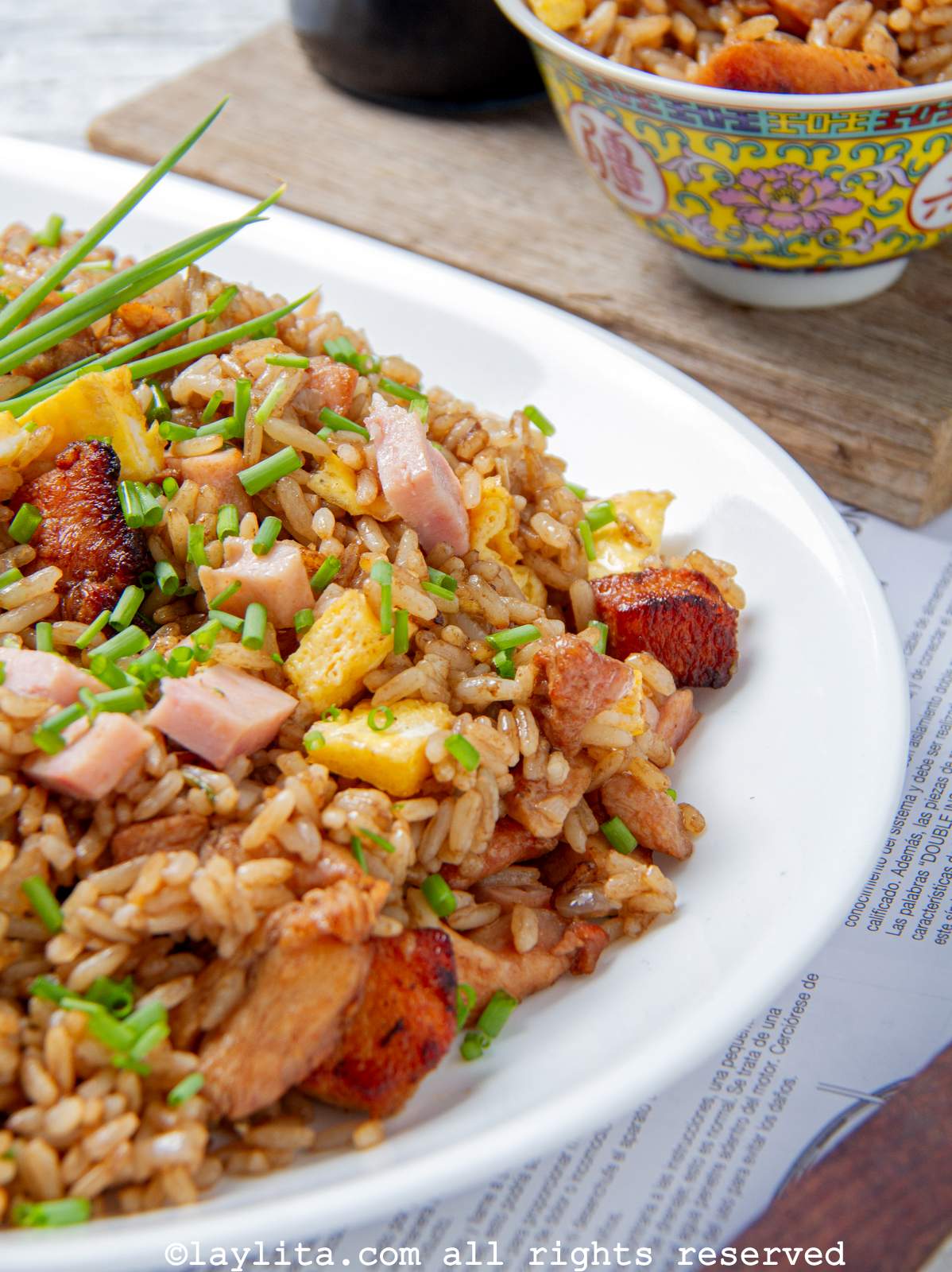 Corner of the serving platter of arroz chaufa with a small bowl of arroz chaufa behind it on a wooden board. 