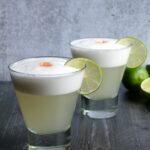 Side view of two pisco sours in tumbler glasses garnished with limes.