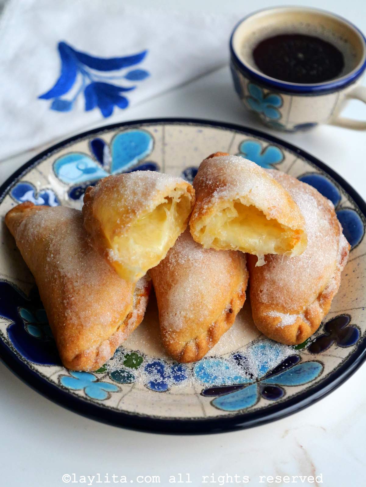 Cheese empanadas on a blue floral plate with one on top cut in half and a cup of coffee in the background