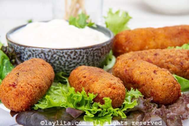 Close up of fried Spanish ham croquettes served on a platter lined with lettuce and a dipping sauce on the side.