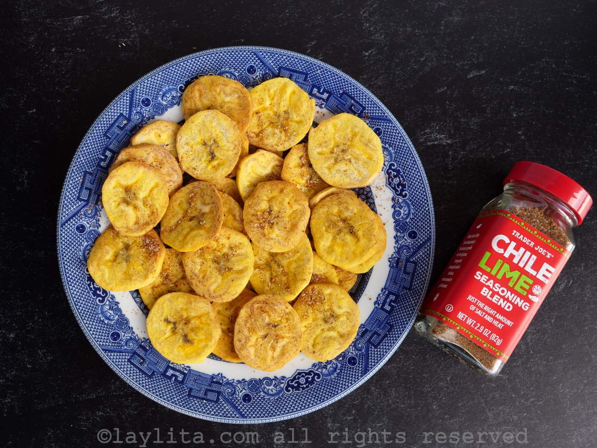 Air fryer plantain chips on a blue plate with a black background next to chili lime salt.