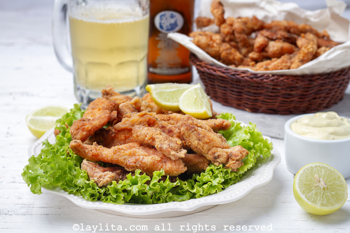 Plate of crispy chicken chicharrones on top of lettuce leaves with lime slices and a mug of beer in the backgroun
