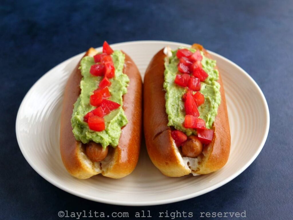 Two hot dogs on a white plate with sausage, avocado and tomatoes on top.