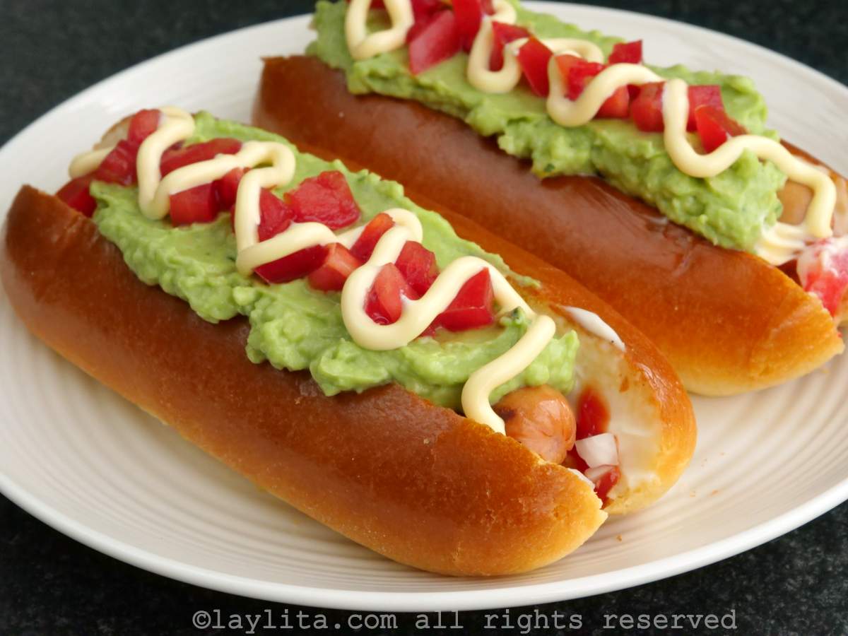 Side view of a chilean completo on a plate with sausage, avocado, tomatoes and a drizzle of mayo on top.
