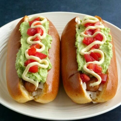 Two Chilean completos on a white plate with tomatoes and mayo on top, with a blue background.