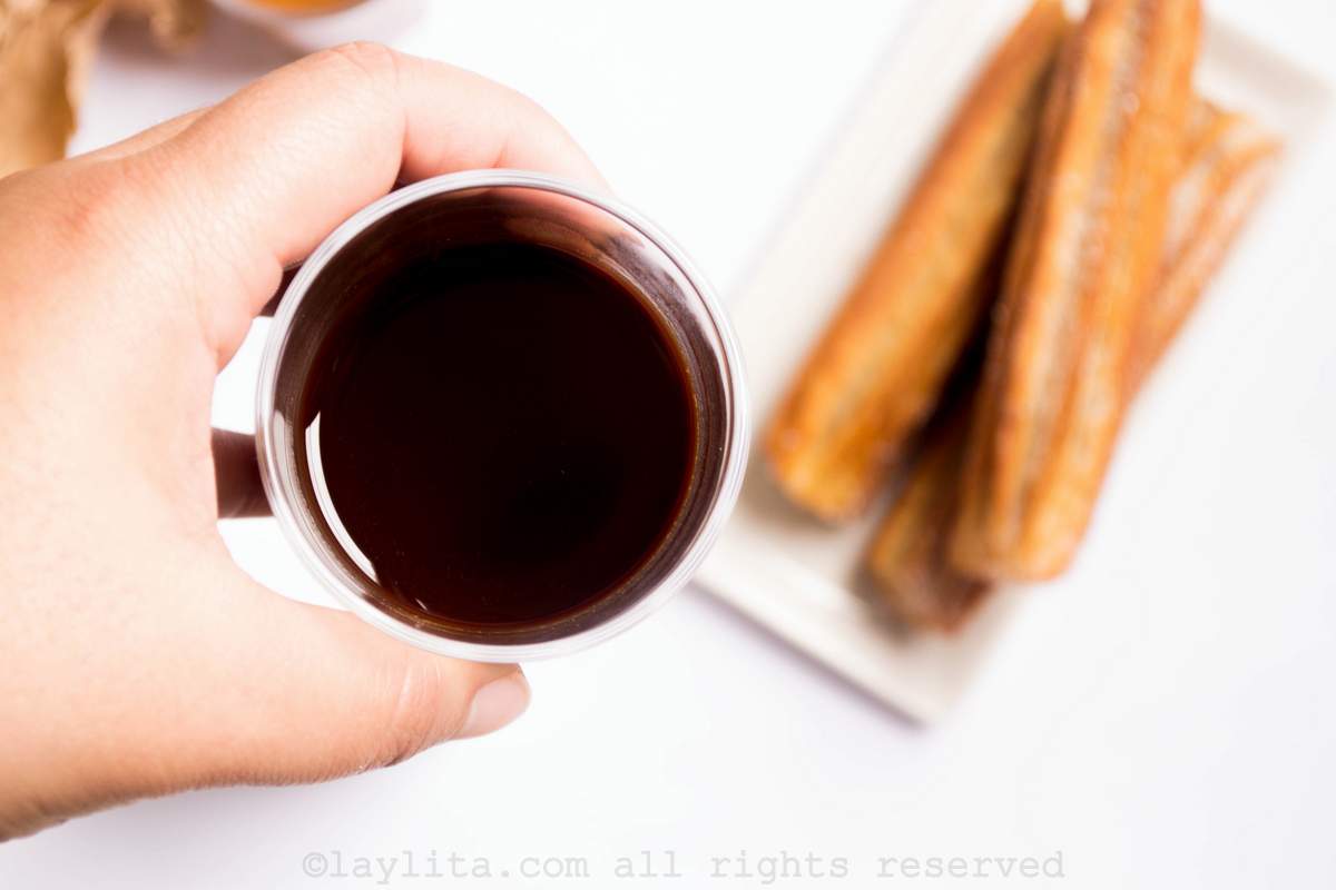 Hand holding a small ramekin of chocolate sauce with a plate of three churros in the background.