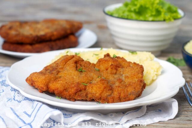 A white plate with a crispy breaded milanesa steak and mashed potatoes, with a salad in the background
