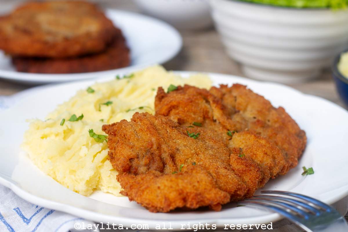 Side view of milanesa de res or thin breaded beef steak with mashed potatoes with a salad and more steaks in the background.