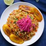White plate of tuna fried rice served with plantains and topped with pickled onions on a blue background.