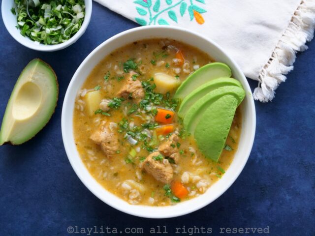 Bowl of sopa de avena con cerdo on a blue background with a small bowl of cilantro and green onions on the side.