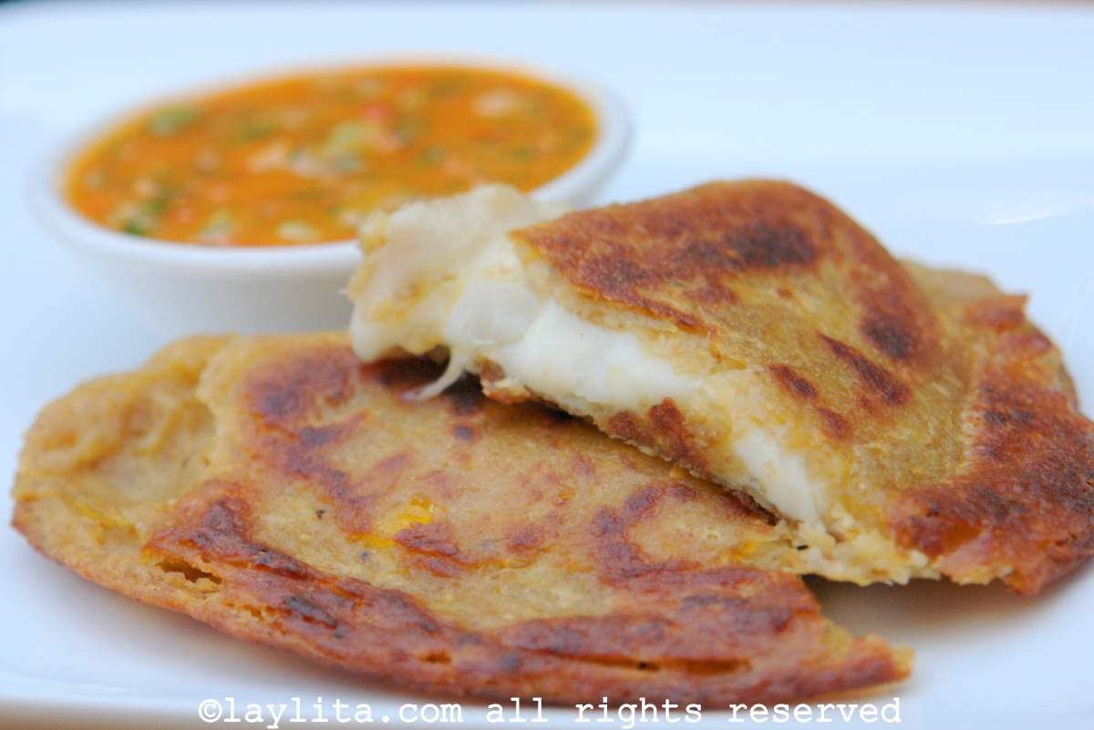 An empanada made with green plantain dough and filled with cheese, cut in half, with cheese oozing out, on a white plate