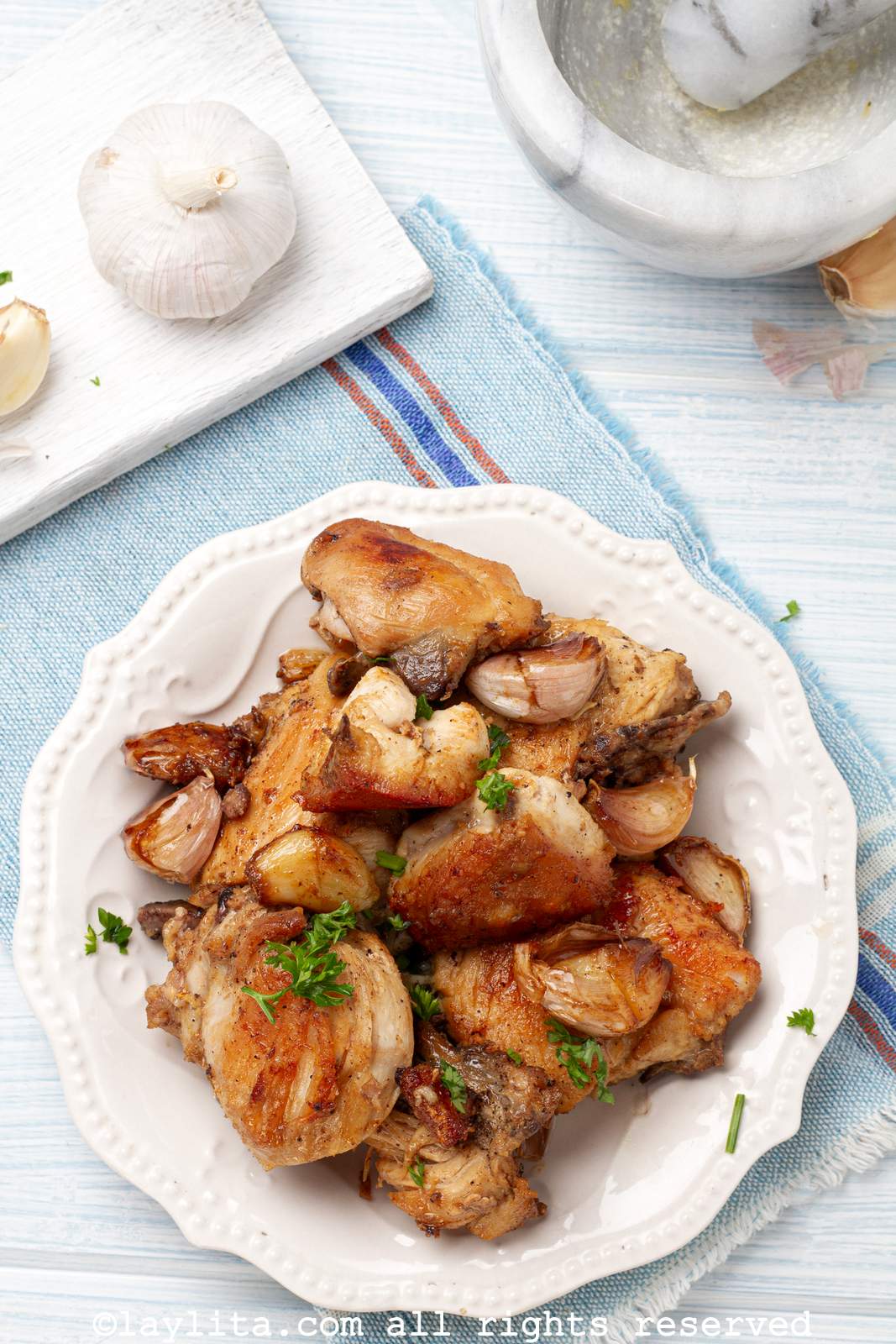 Overhead shot of Spanish garlic chicken on white platter with whole roasted garlic cloves and a whole garlic head in the background