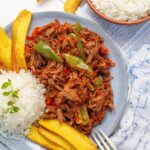 Overhead shot of ropa vieja served on a light blue plate with rice and fried plantains.