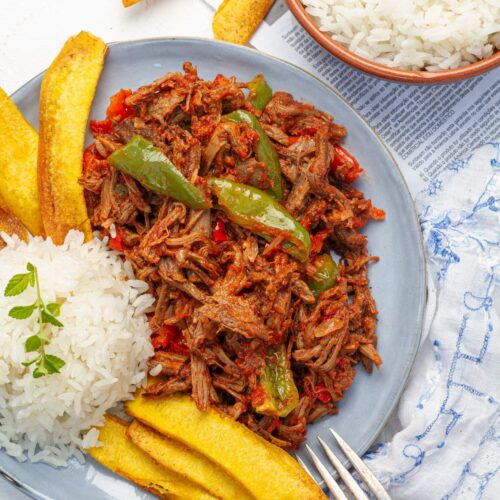 Overhead shot of ropa vieja served on a light blue plate with rice and fried plantains.