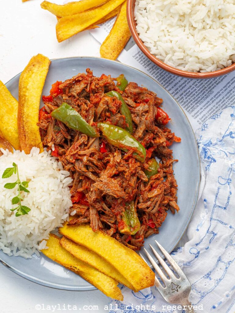 Overhead shot of ropa vieja served on a light blue plate with rice and fried plantains.