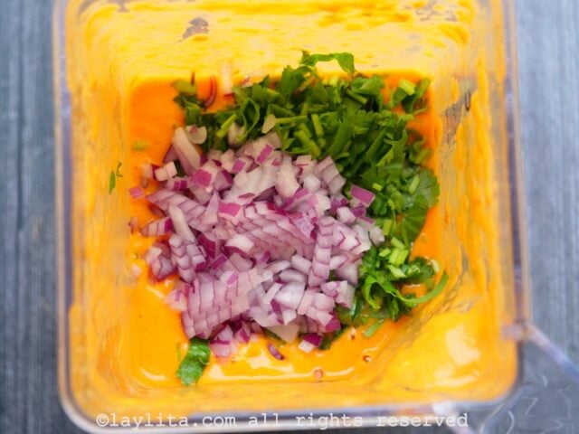 Chopped onions and cilantro being added to a blender with an orange sauce