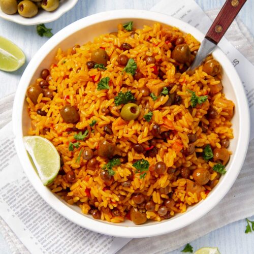 Overhead shot of arroz con gandules in a white bowl with lime wedges on the side and a bowl of olives in the background