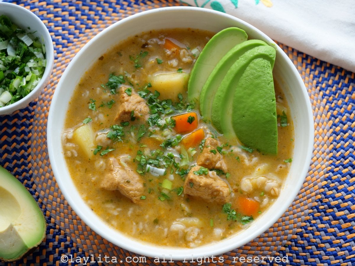 White bowl full of sopa de avena or oatmeal soup with pork and vegetables on a blue and orange placemat served with avocado