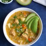 White bowl with oatmeal vegetable pork soup on a blue background with a small bowl of cilantro and green onions on the side.