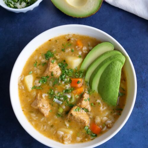 White bowl with oatmeal vegetable pork soup on a blue background with a small bowl of cilantro and green onions on the side.