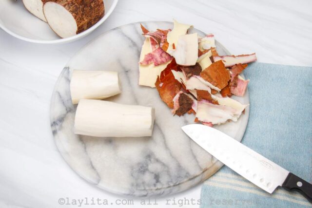 Two pieces of peeled yuca or cassava on a board, next to the peels and a knife