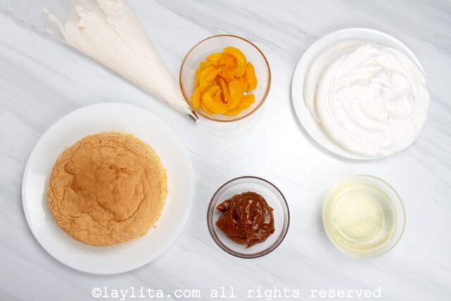 Sponge cake, meringue discs, peaches, dulce de leche and whipped cream on a kitchen counter ready to assemble the Uruguayan Torta Chajá