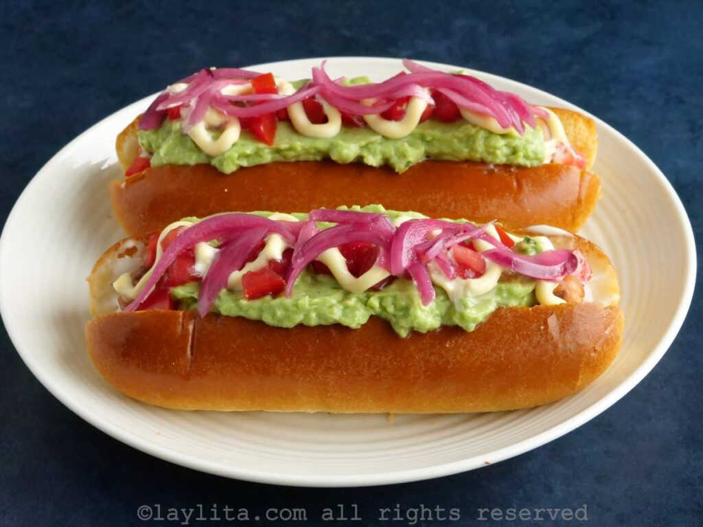 Side view of two Chilean completos on a plate topped with pickled onions.