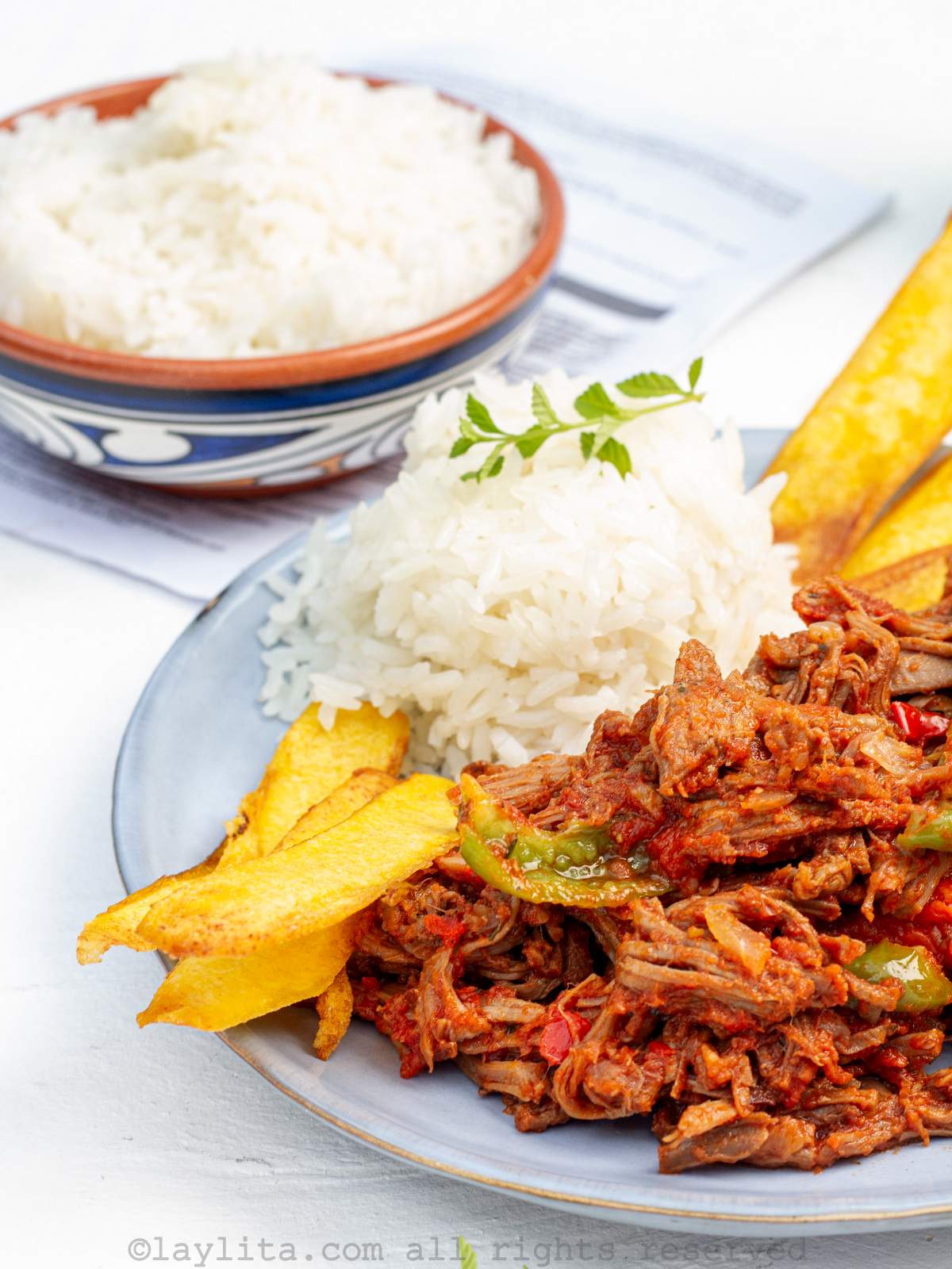 Close up side view of ropa vieja beef on a plate served with rice and plantains with more rice in the background.