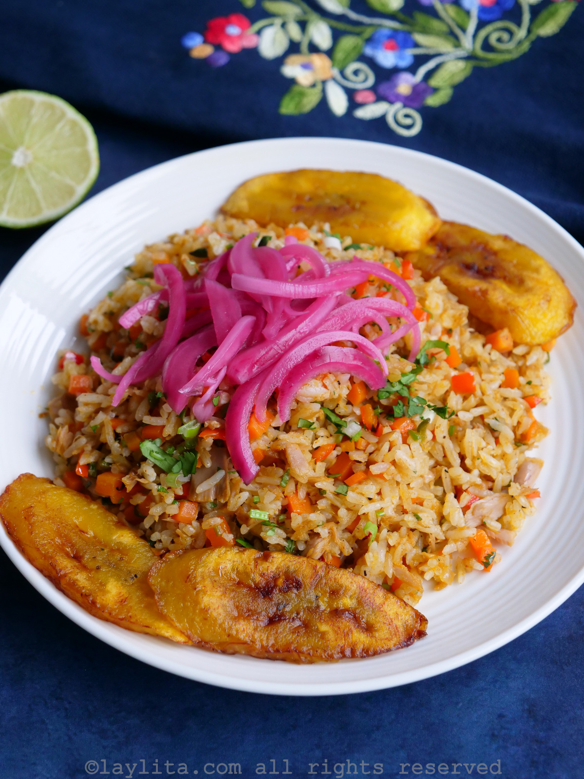 Large serving of arroz con atún on a white plate served with fried plantains and pickled onions, on a blue background.