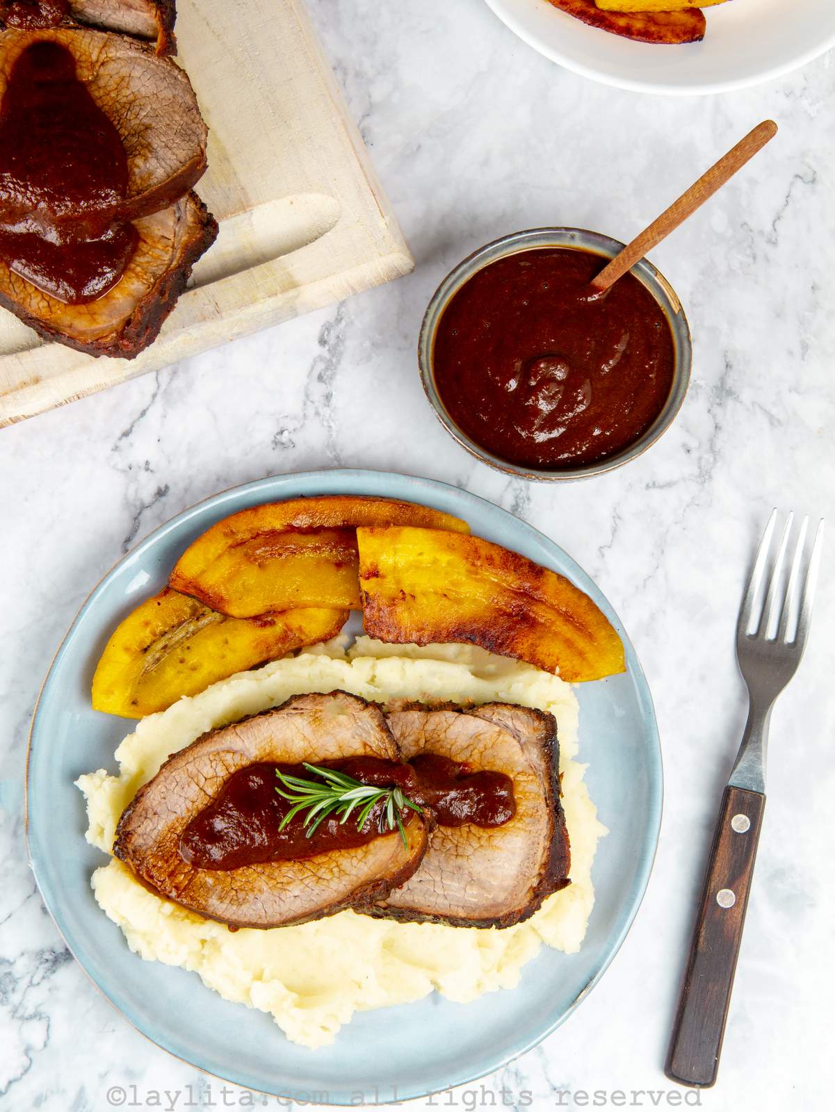 Overhead shot of Venezuelan roasted beef served on mashed potatoes next to sweet plantains topped with sauce with a side of sauce on the side.