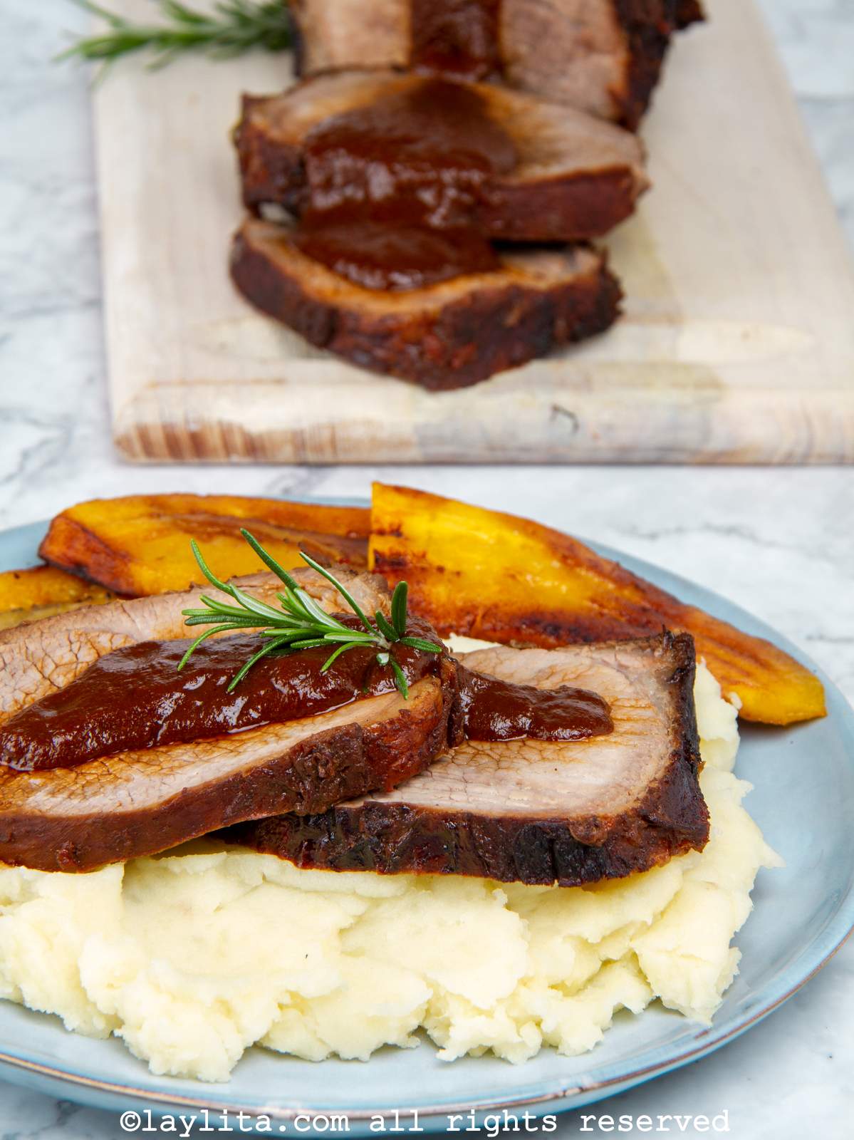 Plate of Venezuelan asado negro with mashed potatoes, plantains and sauce with the rest of the roast in the background.