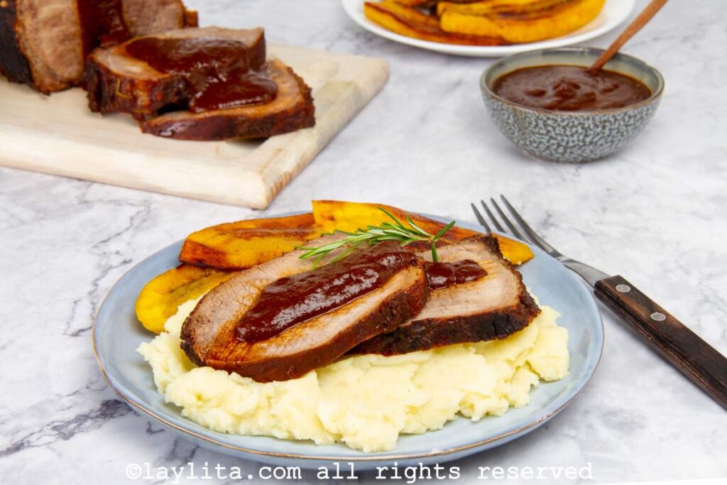Plate of Venezuelan asado negro with mashed potatoes, plantains and sauce with the rest of the roast in the background.