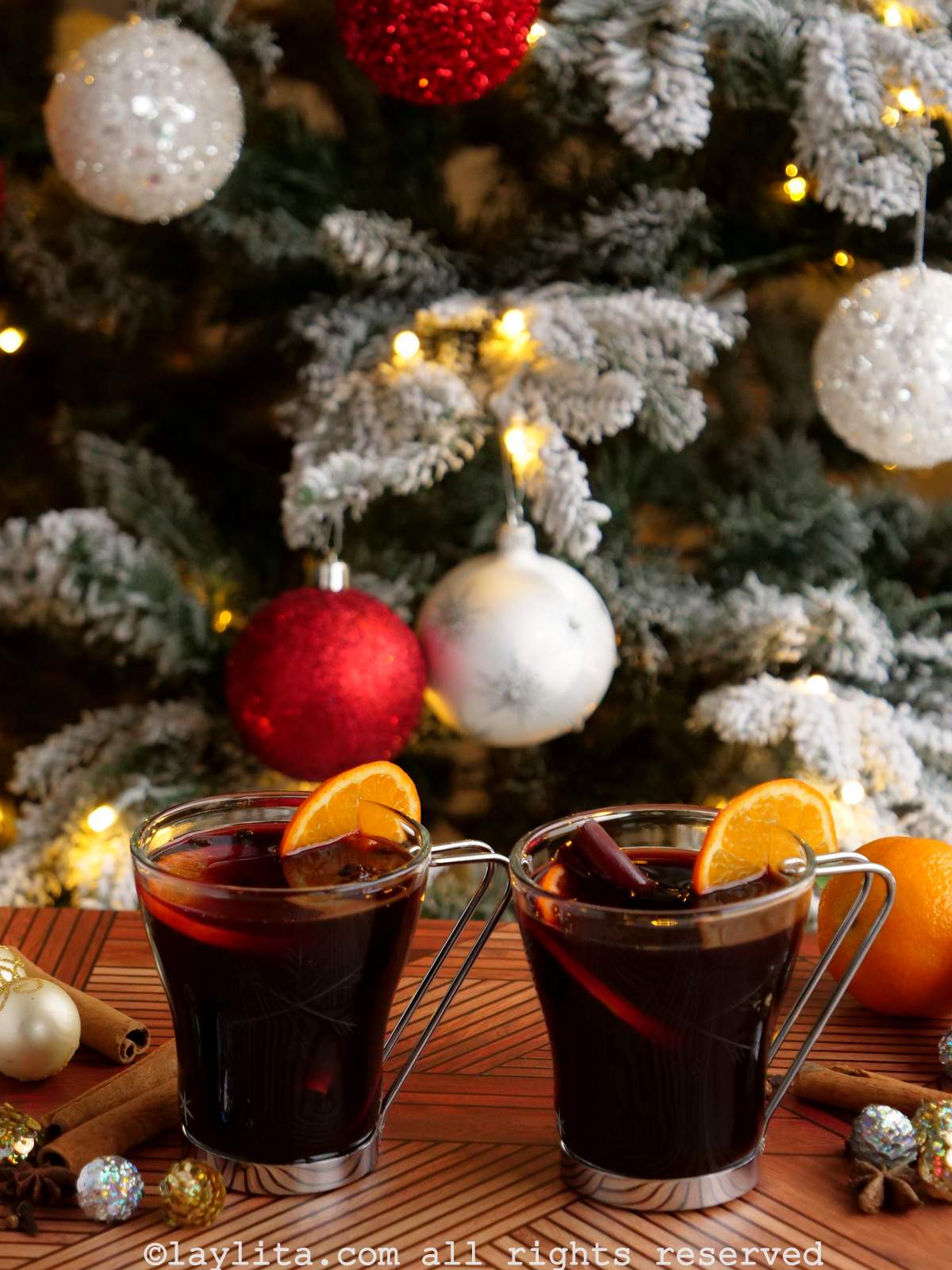 Two glasses of mulled wine garnished with orange slices on a wood table nex to Christmas ornaments and a tree in the background