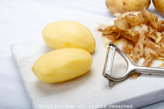 Two peeled potatoes next to a peeler and potato skins