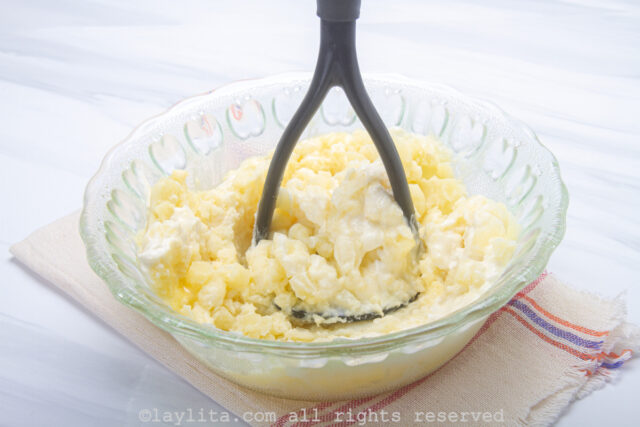 Potatoes being mashed in a glass bowl with a masher