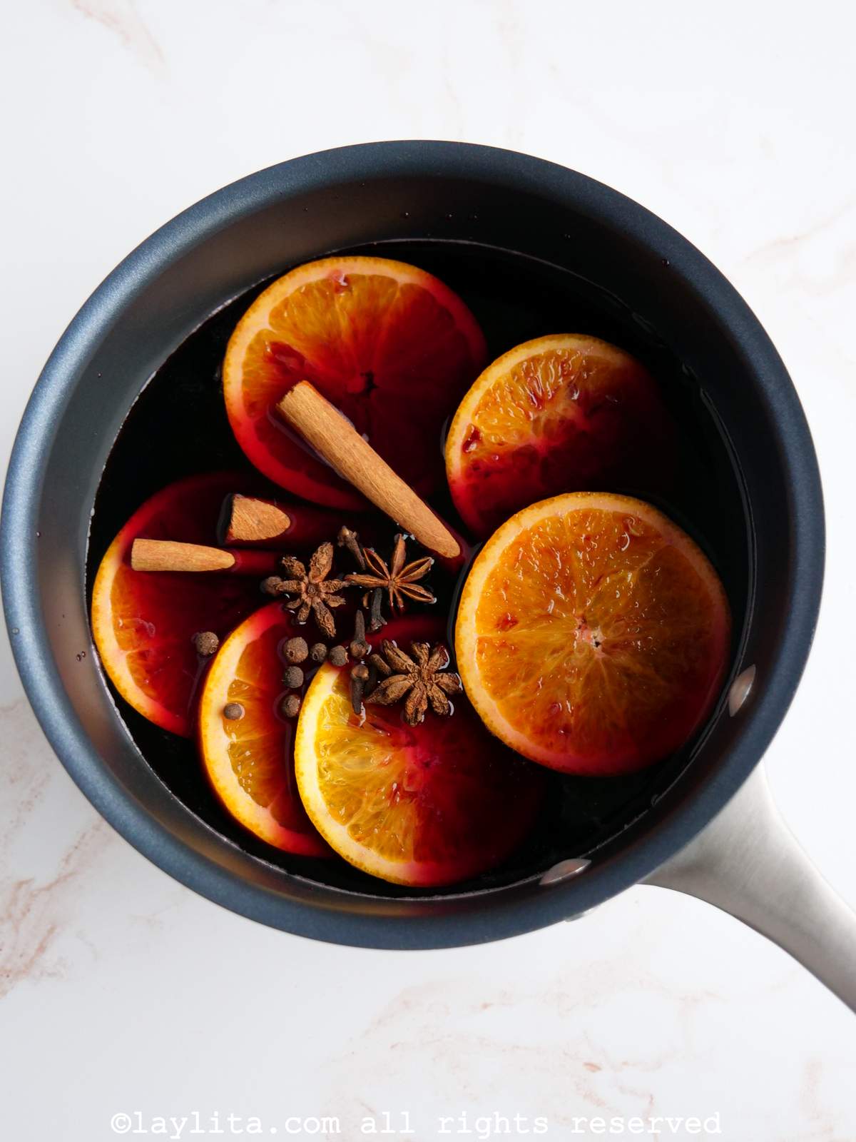 A small saucepan full of red wine, orange slices and spices on a white countertop