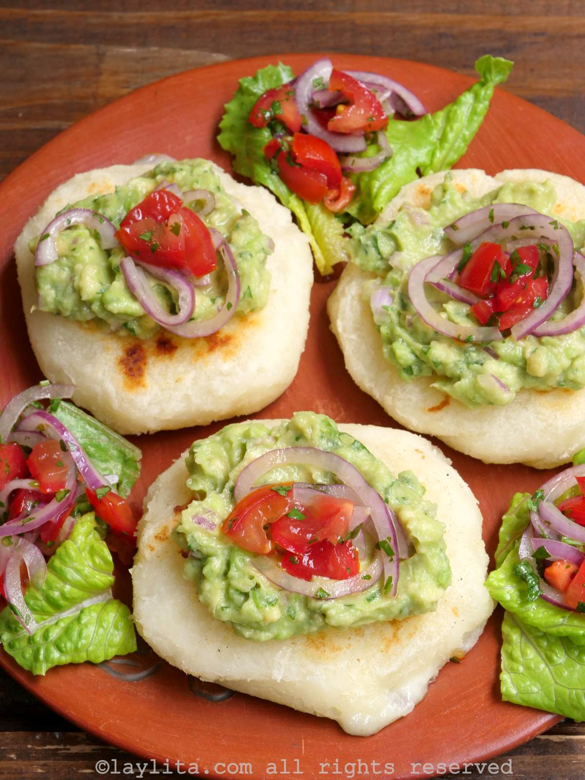 Overhead shot of three tortillas de yuca on a red plate, each topped with avocado and tomato and onion salad.