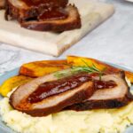 Venezuelan asado negro with mashed potatoes, fried plantains and sauce on a blue plate with the rest of the roast in the background.