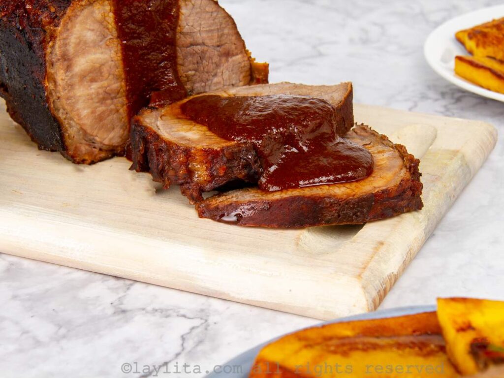 Asado negro served on a wooden board with some slices off the end and a sauce on top, with fried plantains on the side