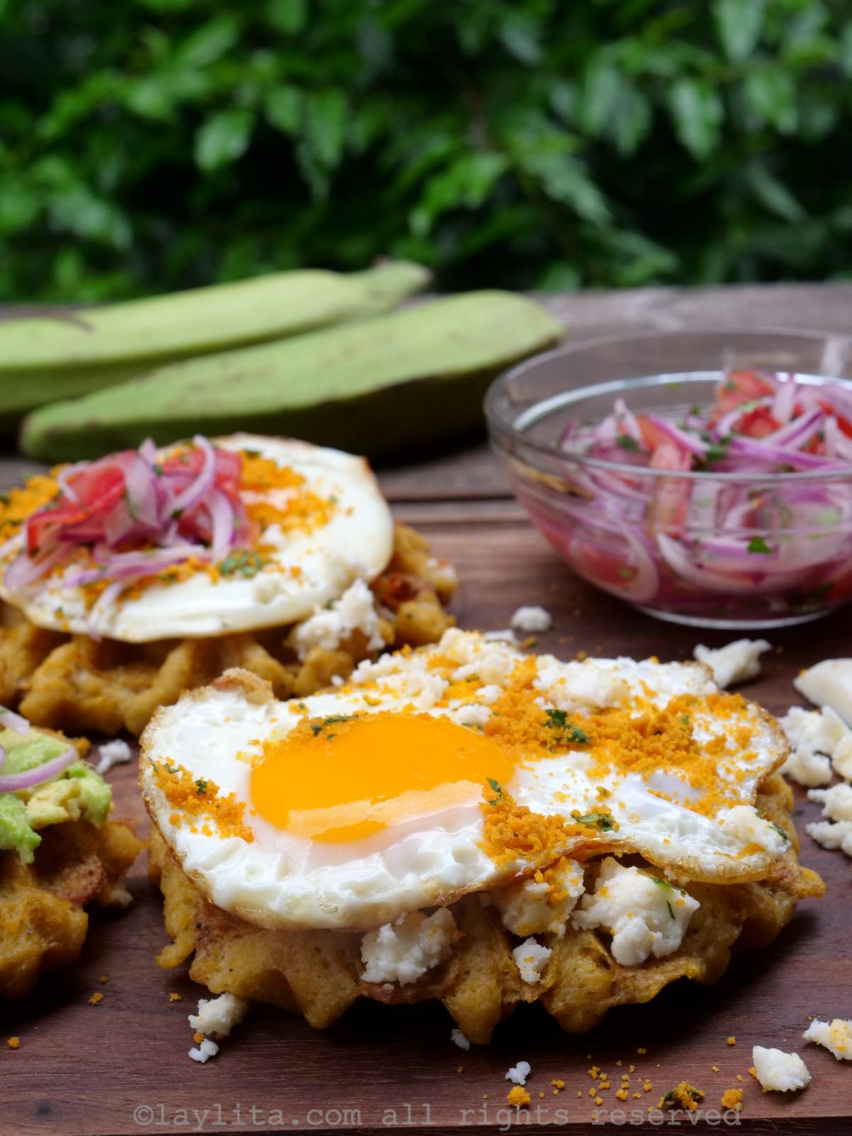 A green plantain waffle topped with cheese and a fried egg, in the background there are more waffles and some green plantains