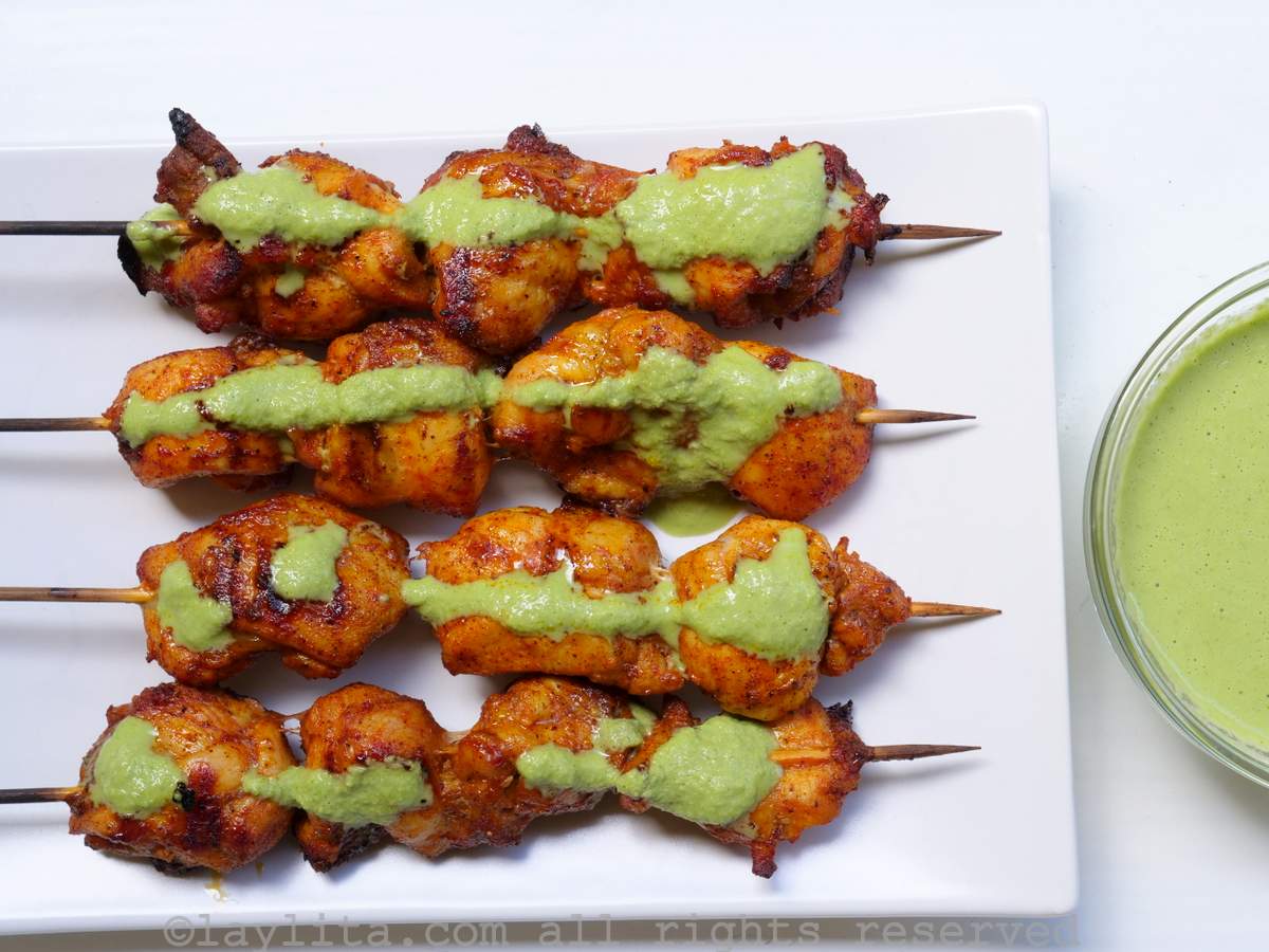 Four chicken skewers on a white plate with a green sauce on top and more sauce on the side.