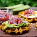 Side view of a green plantain waffle topped with avocado and a tomato onion salad.