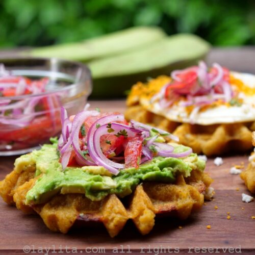 Side view of a green plantain waffle topped with avocado and a tomato onion salad.