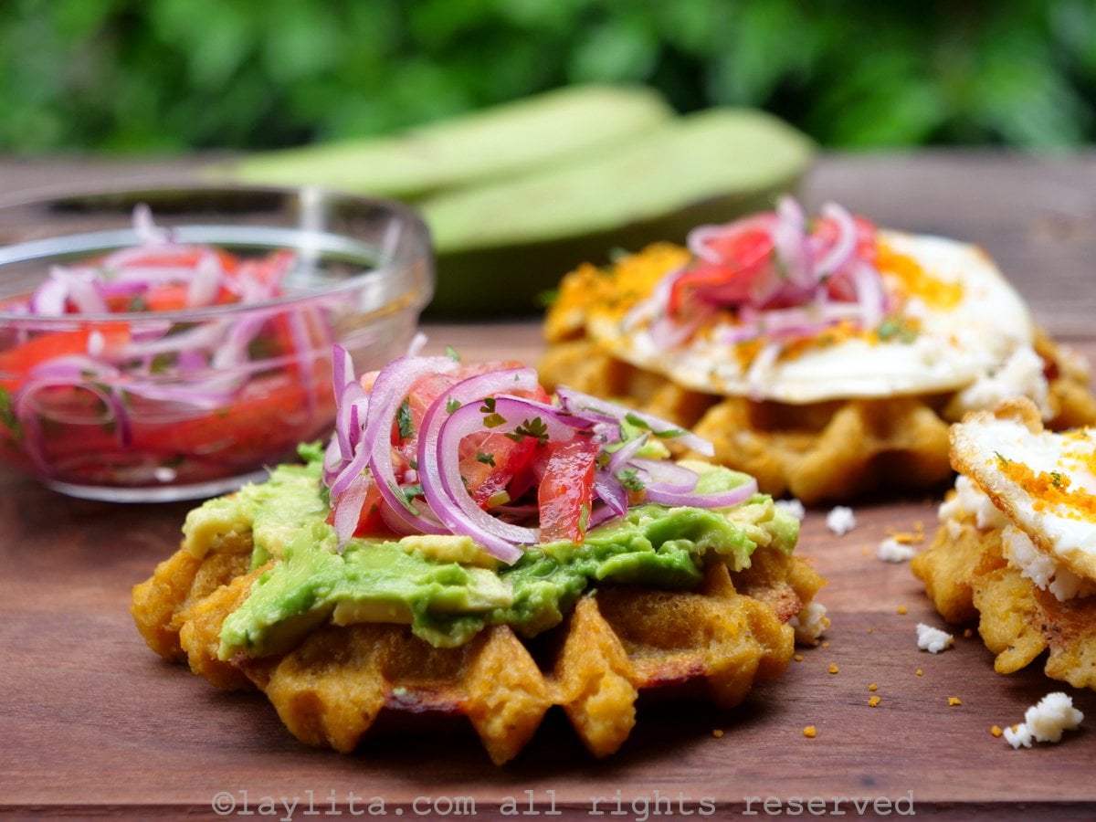 Side view of a green plantain waffle topped with avocado and a tomato onion salad.